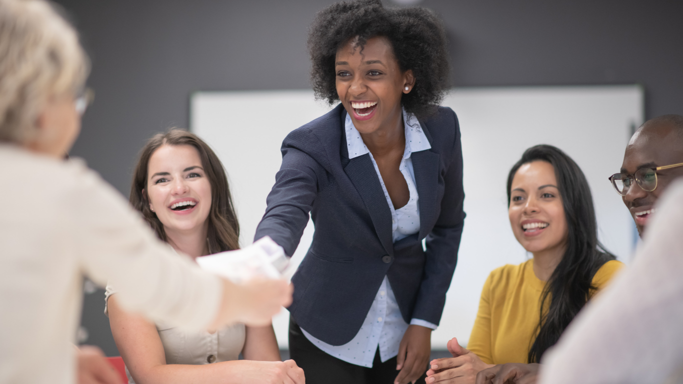 A woman in a suit reaching across the table to a colleague. All participants are smiling broadly.