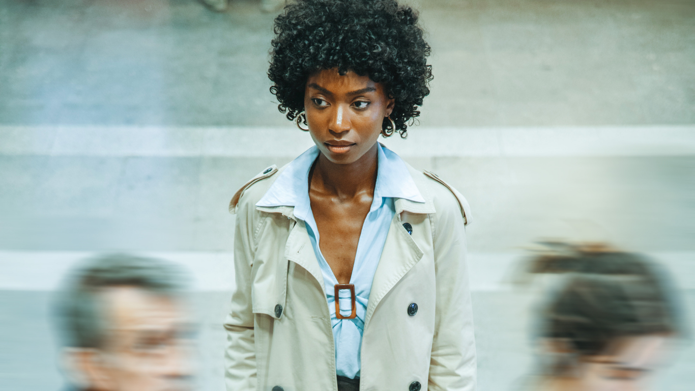 A woman stands still and observant in a busy environment as blurred figures move quickly around her. 