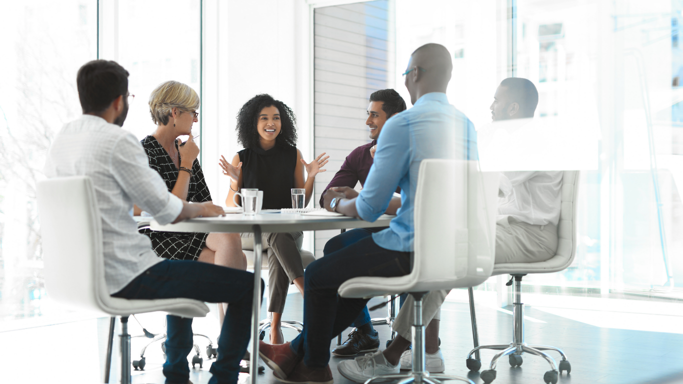 A diverse group of business professionals engaged in a collaborative discussion around a conference table in a bright, modern office. 