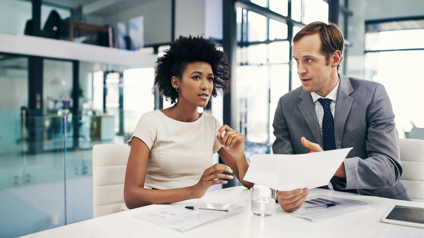 Two business professionals seated at a table discussing a document together in a modern office. 
